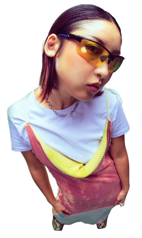 Eine Frau in einem weißen Tshirt mit einem farbenfrohen Kleid darüber und einer Sonnenbrille wird von oben fotografiert. Sie schaut mit leicht geneigtem Kopf in die Kamera.